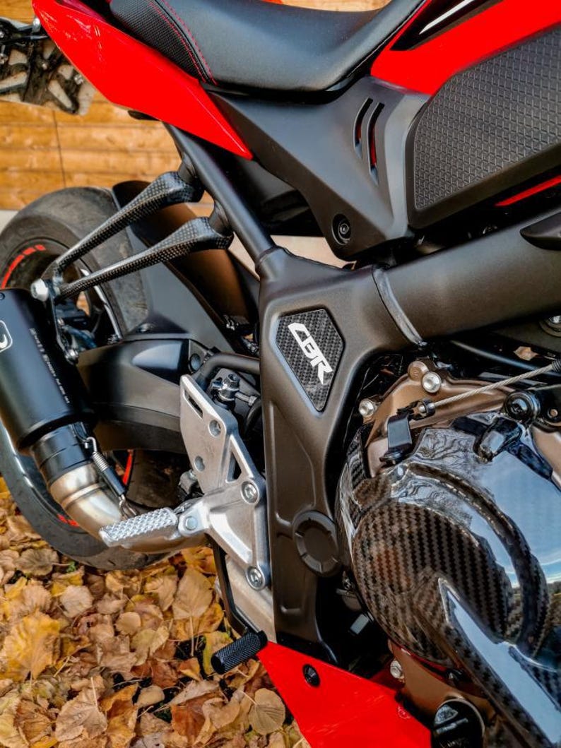 May include: Close-up of a red and black motorcycle with a carbon fiber engine cover. The motorcycle has a black exhaust pipe and a silver footpeg. The word "CBR" is visible on the side of the motorcycle.
