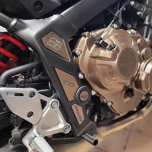 May include: Close-up of a motorcycle engine with a black and brown frame. The engine is made of metal and has a gold finish. The frame has the text "NEO SPORTS CAFE" and "CBR650R" printed on it.