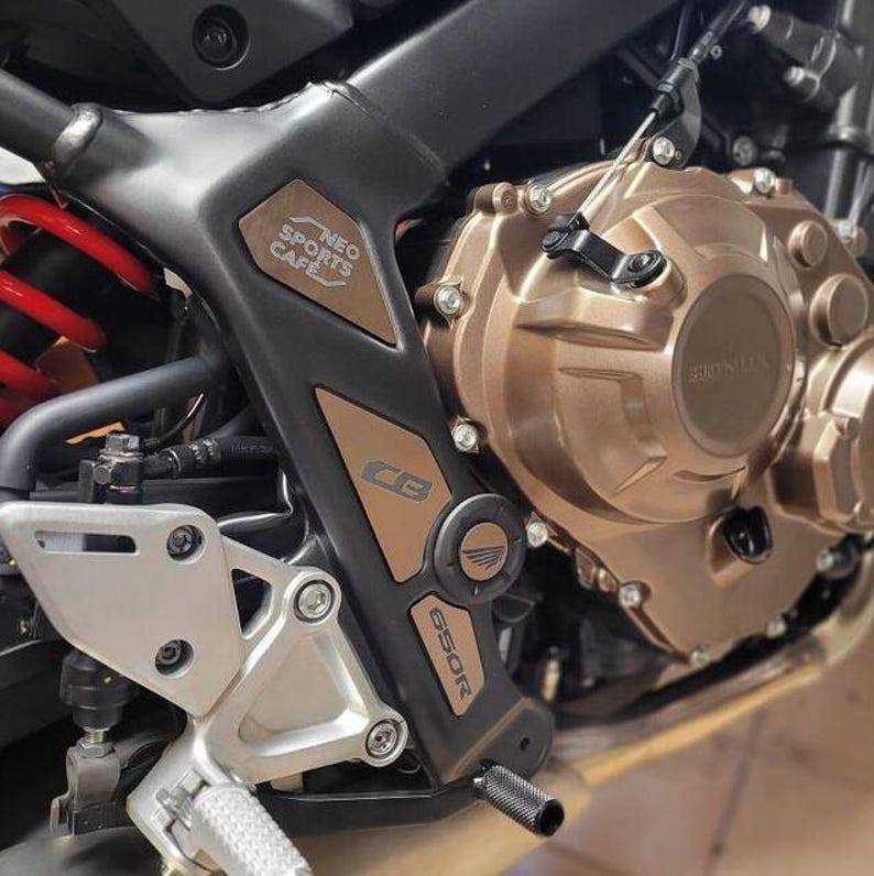May include: Close-up of a motorcycle engine with a black and brown frame. The engine is made of metal and has a gold finish. The frame has the text "NEO SPORTS CAFE" and "CBR650R" printed on it.