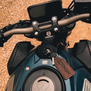 Op de afbeelding: Close-up van een teal motorfiets met zwart stuur en een zwart digitaal display. Een bruin leren label met het woord "ZORKYX" hangt aan de sleutel. De motorfiets heeft een zwarte brandstoftank en een zilveren tankdop.