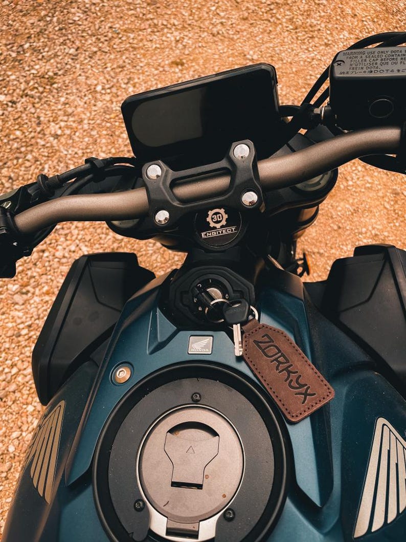 Op de afbeelding: Close-up van een teal motorfiets met zwart stuur en een zwart digitaal display. Een bruin leren label met het woord "ZORKYX" hangt aan de sleutel. De motorfiets heeft een zwarte brandstoftank en een zilveren tankdop.