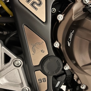 May include: Close-up of a motorcycle engine with two metal plates. The top plate is a triangular shape with the number "12" in black lettering. The bottom plate is rectangular with the number "98" in black lettering. The plates are attached to the engine with screws.