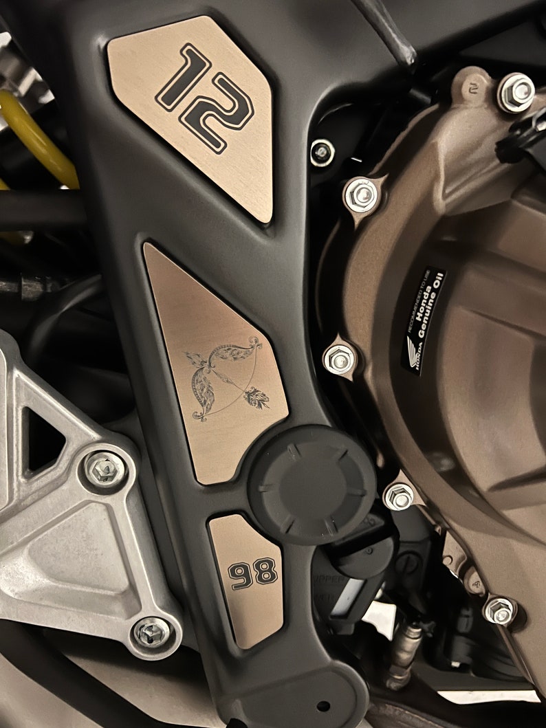 May include: Close-up of a motorcycle engine with two metal plates. The top plate is a triangular shape with the number "12" in black lettering. The bottom plate is rectangular with the number "98" in black lettering. The plates are attached to the engine with screws.