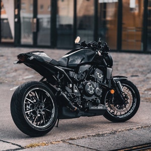 Può includere: Una motocicletta nera con uno scarico personalizzato e ruote nere. La motocicletta è parcheggiata su una superficie di cemento grigia.