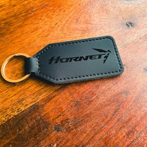 May include: Black rectangular keychain with the word "Hornet" and a stylized graphic. The keychain has a gold-coloured ring and is made of a dark material with stitched edges. The background is a wooden surface.
