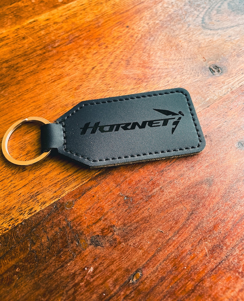 Op de afbeelding: Zwarte rechthoekige sleutelhanger met het woord "Hornet" en een gestileerde afbeelding. De sleutelhanger heeft een goudkleurige ring en is gemaakt van een donker materiaal met gestikte randen. De achtergrond is een houten oppervlak.