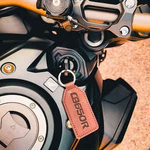 Op de afbeelding: Close-up van het stuur en het contactslot van een motorfiets. Een bruine leren sleutelhanger met de tekst "CB650R" hangt aan het contactslot. De motorfiets heeft zwarte en gouden accenten.