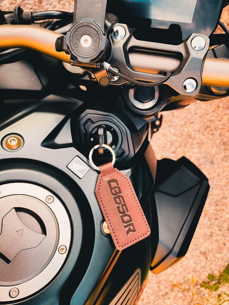 Op de afbeelding: Close-up van het stuur en het contactslot van een motorfiets. Een bruine leren sleutelhanger met de tekst "CB650R" hangt aan het contactslot. De motorfiets heeft zwarte en gouden accenten.