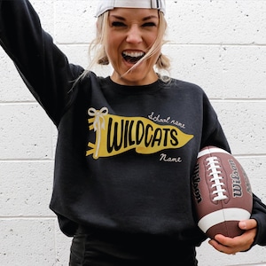 May include: A black long-sleeve crewneck sweatshirt featuring a yellow pennant graphic with a white bow. The pennant displays the word WILDCATS in black lettering, with the text School name and Name printed in white script above and below it.