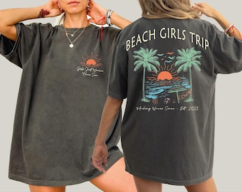 May include: Dark gray t-shirt with a beach-themed graphic. The front features a sun graphic and the text "Girls Just Wanna Have Sun." The back displays "Beach Girls Trip" above a beach scene with palm trees, a sunset, and the text "Making Waves Since - Est. 2023."