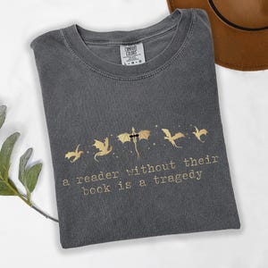 May include: A charcoal gray crew neck t-shirt featuring a gold graphic of five flying dragons and the text "a reader without their book is a tragedy" printed in a typewriter-style font. The shirt has a Comfort Colors brand label at the neck.