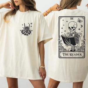 two women wearing white t-shirts with black designs. The t-shirt on the left has a graphic of a skeleton holding a book, while the t-shirt on the right features a tarot card design with a skeleton reading the "Reader" card.