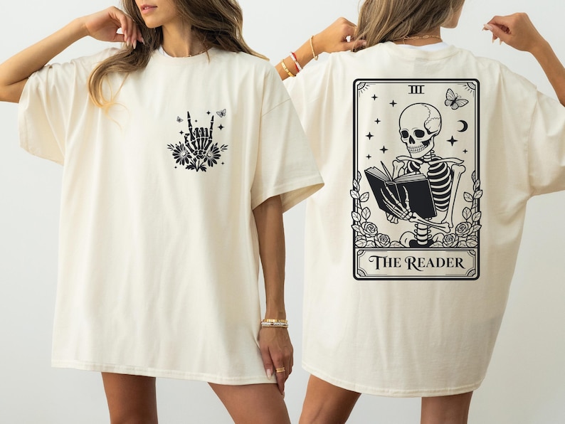 two women wearing white t-shirts with black designs. The t-shirt on the left has a graphic of a skeleton holding a book, while the t-shirt on the right features a tarot card design with a skeleton reading the "Reader" card.