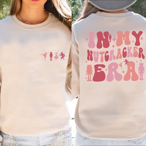 May include: Cream-colored sweatshirt with a ballet-themed design. The front features a row of ballet figures in pink. The back displays the text "IN MY NUTCRACKER ERA" in shades of pink and red, along with nutcracker and ballerina illustrations.