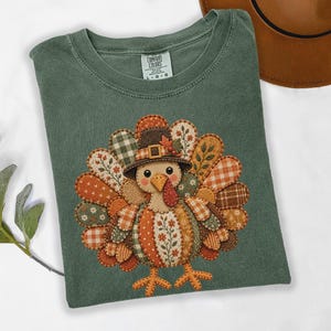 May include: A sage green t-shirt featuring a graphic of a turkey wearing a pilgrim hat. The turkey is composed of various patterned patches in shades of orange, brown, and cream, including floral, plaid, and gingham designs. The shirt has a Comfort Colors label.