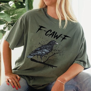May include: Olive green t-shirt featuring a black raven perched on a branch, with the text "F-CAW-F" above. The design includes splatter details. The shirt is a casual, short-sleeved style.