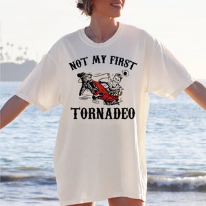 May include: White t-shirt with a black graphic of a person riding in a red car being lifted by a tornado. The text "NOT MY FIRST TORNADEO" is printed on the shirt.