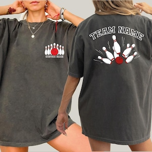 May include: Dark gray t-shirt with a bowling theme. The front features a graphic of bowling pins and a red ball, with the text "CUSTOM MADE". The back displays a bowling strike graphic with the text "TEAM NAME".