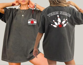 May include: Dark gray t-shirt with a bowling theme. The front features a graphic of bowling pins and a red ball, with the text "CUSTOM MADE". The back displays a bowling strike graphic with the text "TEAM NAME".