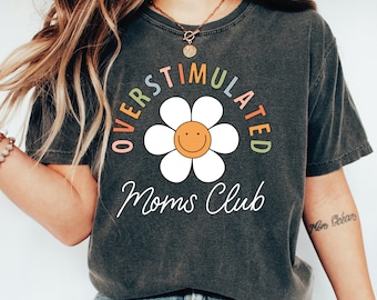 May include: A dark gray t-shirt with a white daisy and the text "Overstimulated Moms Club" in a rainbow arc around the flower.
