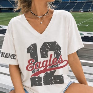 May include: A white V-neck t-shirt featuring a large, distressed dark gray number 12 graphic on the front, with the word Eagles in red script layered over it. The right sleeve displays the word NAME in a distressed dark gray font.
