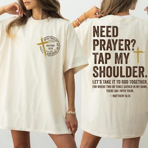 May include: Cream-colored t-shirt with the text "NEED PRAYER? TAP MY SHOULDER" and a cross graphic. The back of the shirt has the same text and a Bible verse. A casual, comfortable tee.