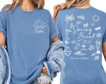 Op de afbeelding: Blauw t-shirt met een witte afbeelding van een zon boven water en het woord "Custom" op de voorkant. De achterkant toont "Spring Break 2026" met strandillustraties en "Cancun Mexico".