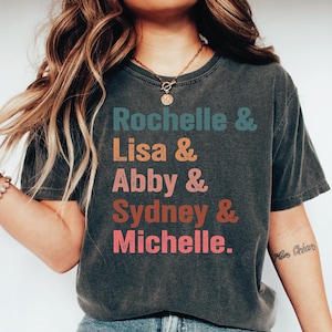 May include: Dark gray t-shirt with the names Rochelle, Lisa, Abby, Sydney, and Michelle printed in teal, orange, pink, brown, and red. The shirt has a vintage, worn look.