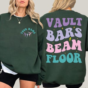 May include: Dark green sweatshirt with the words "VAULT BARS BEAM FLOOR" in purple, light purple, pink, and teal on the back. The front has the text "YOUR NAME" and a gymnast graphic. The sweatshirt is a comfortable, casual style.