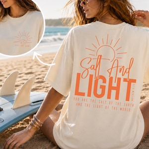 May include: An off-white short-sleeve t-shirt featuring orange graphics. The back displays a sun icon, the text Salt And Light, and the verse Matthew 5:13-14. The front chest area features a smaller matching sun and wave graphic with the text Salt And Light.
