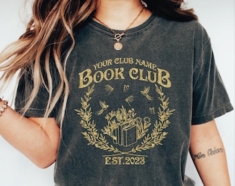 May include: Dark gray t-shirt with a gold design that reads "Your Club Name Book Club" with a floral wreath, books, and the text "EST.2023". The shirt is a crew neck and has short sleeves.