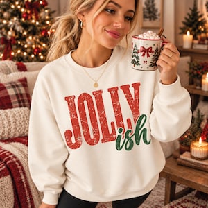 Puede incluir: Una sudadera de cuello redondo color crema con el texto JOLLY en letras rojas desgastadas e ish en letras verdes desgastadas. La prenda tiene mangas largas y cuello, puños y dobladillo acanalados, lo que proporciona un ajuste relajado.