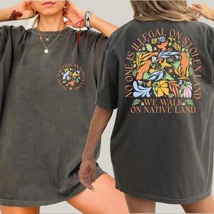 May include: A dark gray oversized t-shirt with a graphic print on the back that says "No one is illegal on stolen land. We walk on native land." The graphic features colorful flowers and birds.