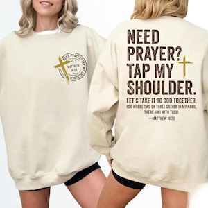 May include: Cream-colored sweatshirt with the text "NEED PRAYER? TAP MY SHOULDER" in brown. The front features a cross and the text "NEED PRAYER? TAP MY SHOULDER" and "Matthew 18:20". The back has a longer quote from Matthew 18:20.
