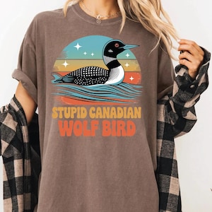 May include: Brown t-shirt featuring a graphic of a loon swimming on water, set against a sunset backdrop. The text "STUPID CANADIAN WOLF BIRD" is printed below the graphic in orange and yellow.