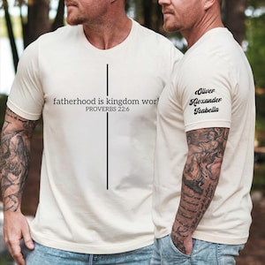 May include: Cream-colored t-shirt with the text "fatherhood is kingdom wor!" and "PROVERBS 22:6" printed in black. A vertical black line is centered on the shirt. The sleeve has the names "Oliver Alexander Isabella" printed in black.