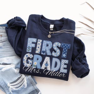 May include: A navy blue crewneck sweatshirt featuring the text FIRST GRADE in large, appliqued letters with various blue and white floral, gingham, and polka dot patterns. The name Mrs. Miller is printed in white cursive script below the text.