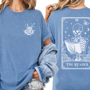 A woman wearing a blue t-shirt with a graphic design on the back, depicting a skeleton reading a book.