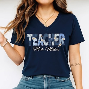 May include: A navy blue V-neck t-shirt featuring the word TEACHER in large, blue-toned patterned block letters decorated with small white daisy flowers. Below the word, the text Mrs. Miller is printed in a white, cursive script font.