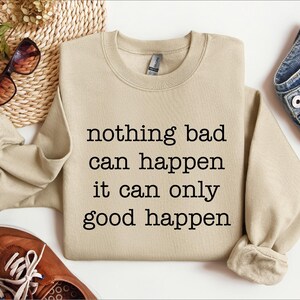 a beige sweatshirt with the text "nothing bad can happen it can only good happen" printed on it, along with a pair of sunglasses and a pair of jeans.