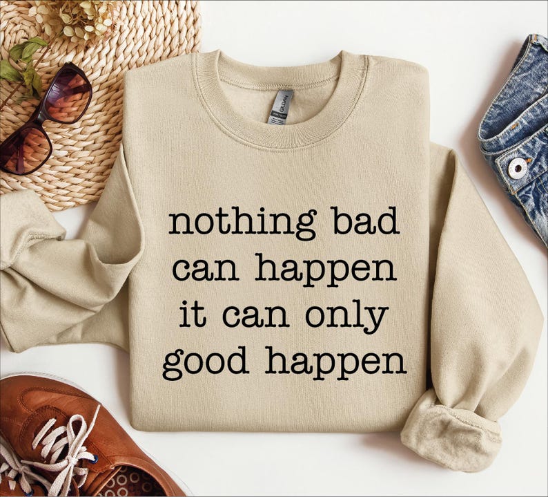 a beige sweatshirt with the text "nothing bad can happen it can only good happen" printed on it, along with a pair of sunglasses and a pair of jeans.