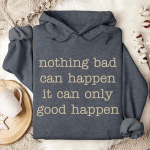 A gray hoodie with the text "nothing bad can happen, it can only good happen" printed on it, along with a mug and some white flowers on a bed.