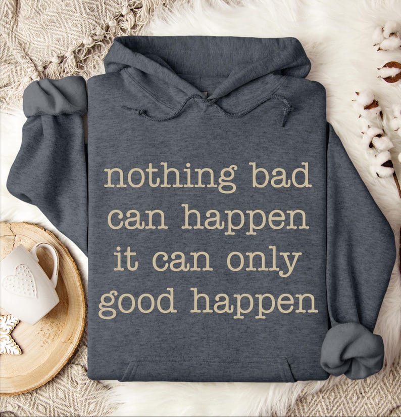 A gray hoodie with the text "nothing bad can happen, it can only good happen" printed on it, along with a mug and some white flowers on a bed.