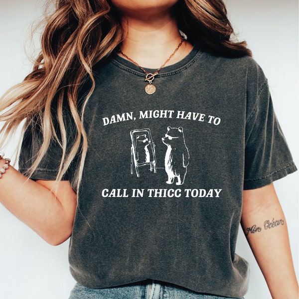 Puede incluir: Una camiseta gris oscuro con un gráfico blanco de un oso con gafas de sol mirando en un espejo. El texto en la camiseta dice "Damn, might have to call in thicc today."