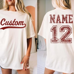 Puede incluir: Camiseta color crema con la palabra "Custom" en escritura burdeos en la parte delantera. La parte trasera de la camiseta muestra la palabra "NAME" encima del número "12" en una fuente burdeos a juego, de estilo universitario. La camiseta tiene un corte holgado.