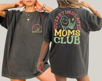 May include: Dark gray t-shirt with the text "OVERSTIMULATED MOMS CLUB" in a colorful design. The shirt features a graphic of a melting smiley face, a lightning bolt, and a skeleton hand.
