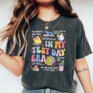May include: Dark gray t-shirt with the text "IN MY TEST DAY ERA" in colorful, retro-style lettering. The shirt features various school-related graphics, including a book, ruler, clock, and pencils. Additional text includes "You know it, now show it" and "You got this."