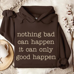 a brown hoodie with the text "nothing bad can happen it can only good happen" printed on it, along with a white mug and some white flowers in the background.
