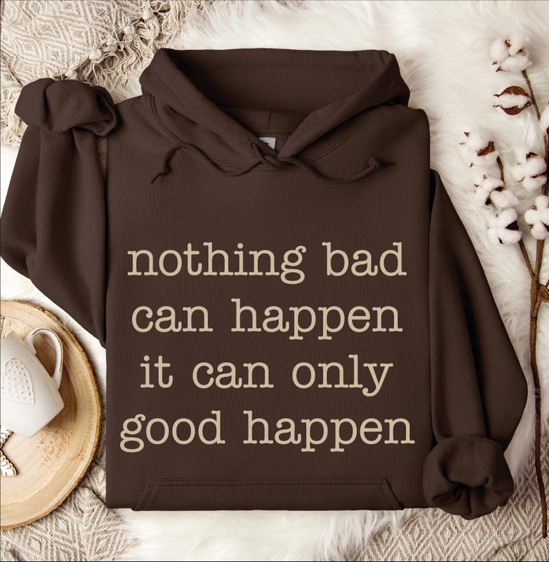 a brown hoodie with the text "nothing bad can happen it can only good happen" printed on it, along with a white mug and some white flowers in the background.