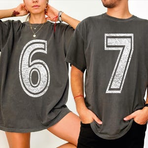 May include: Two charcoal gray t-shirts, each with a large, distressed white number. One shirt displays the number "6", the other displays "7". The shirts have short sleeves and a relaxed fit. The image shows the shirts being worn by two people.